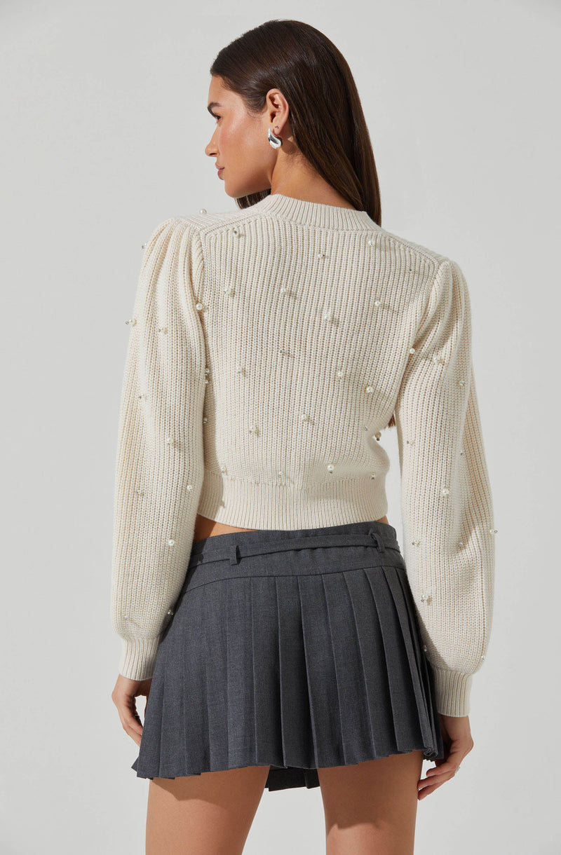 A woman is wearing a cream-colored knit sweater with pearl embellishments on the sleeves and back. The sweater has a ribbed neckline, cuffs, and hem. She pairs it with a high-waisted, pleated, dark gray skirt.