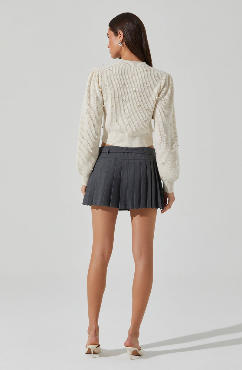 A woman is wearing a cream-colored, long-sleeved, knitted sweater with small pearl-like embellishments throughout. She pairs it with a high-waisted, dark gray pleated mini skirt, and white high-heeled shoes.