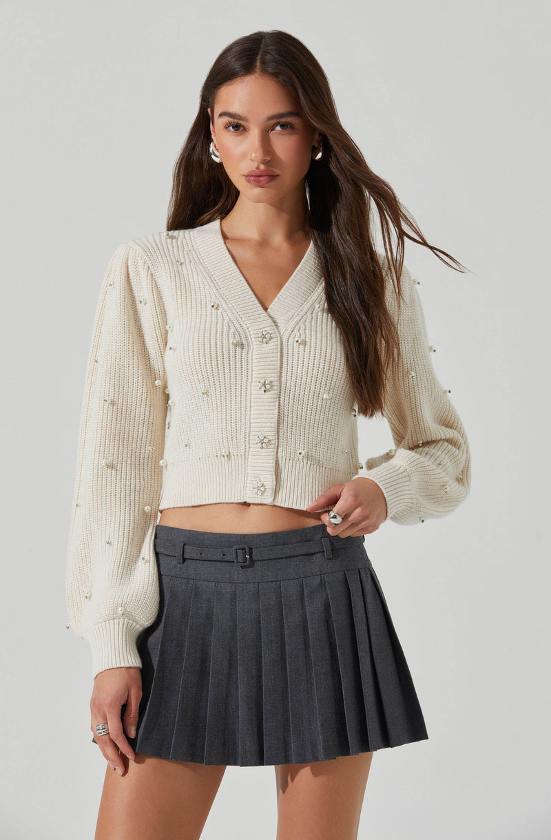 The woman is wearing a cream-colored, cropped cardigan sweater with a V-neckline and long, slightly puffed sleeves. The cardigan has decorative pearl embellishments and button closures down the front. She pairs it with a high-waisted, dark gray pleated mini skirt featuring a waistband and belt loops.