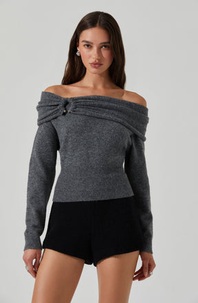 A woman is wearing an off-the-shoulder, long-sleeved, dark gray sweater with a textured, draped detail on the left shoulder, secured with a small button. She is also wearing black shorts and accessorized with earrings and rings.