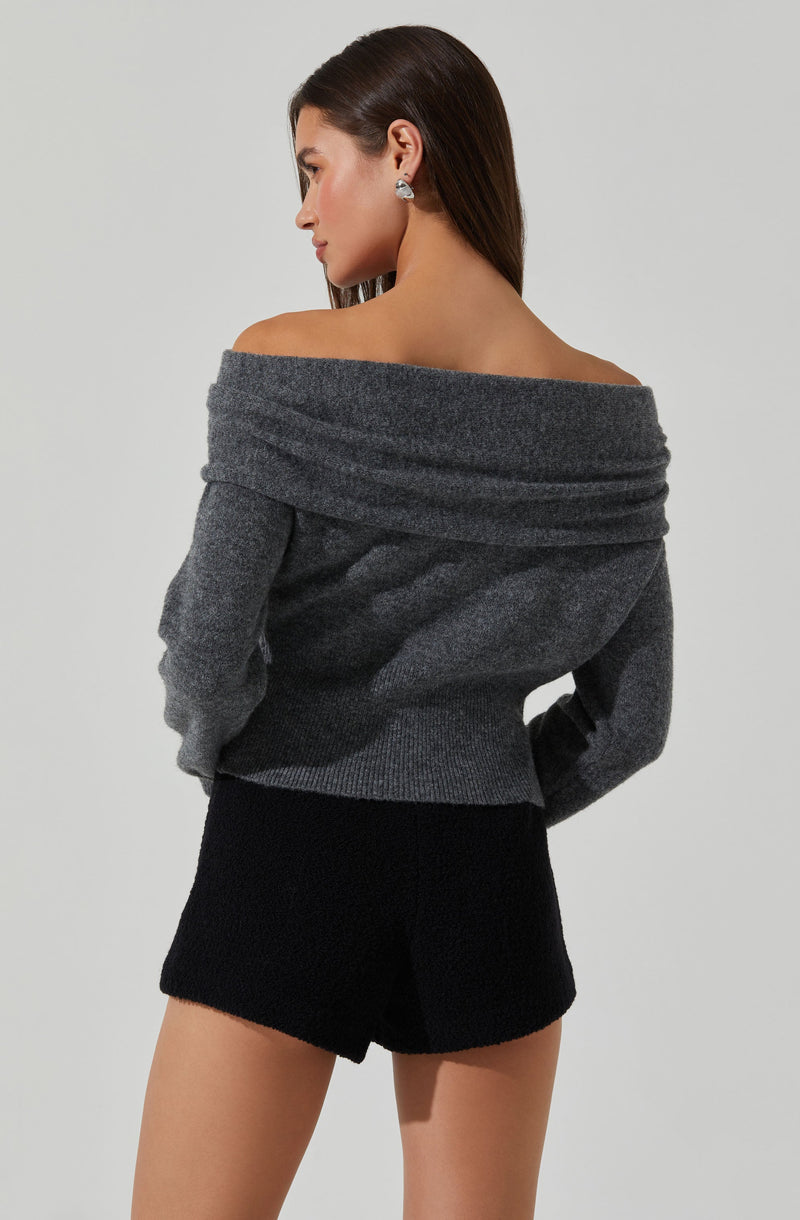 A woman wearing an off-the-shoulder, long-sleeved, gray sweater with a wide neckline that drapes over her shoulders. She has on small, sparkly stud earrings. She is also wearing black shorts with a textured fabric.