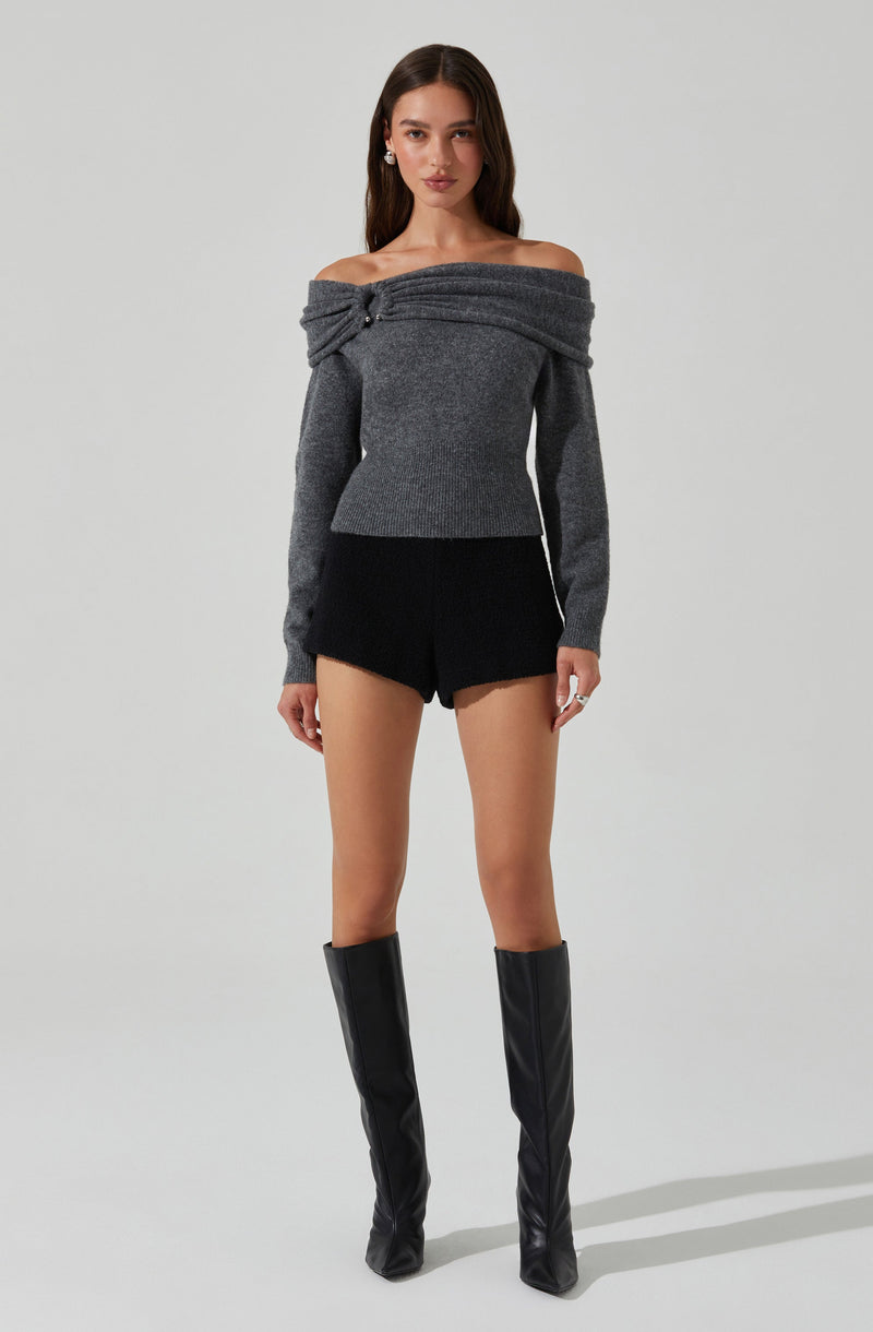 A woman is wearing an off-the-shoulder, long-sleeved gray sweater with a knot detail at the shoulder. She pairs it with black shorts and tall, black knee-high boots.