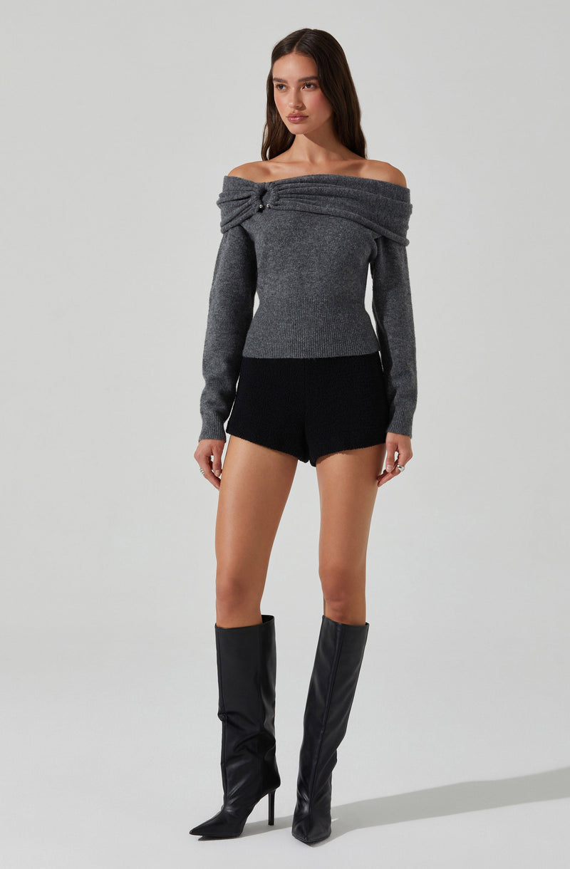 A woman is wearing an off-the-shoulder, long-sleeved, gray sweater with a wrapped and buttoned detail at the shoulder. She pairs it with black high-waisted shorts and tall black leather boots with stiletto heels.