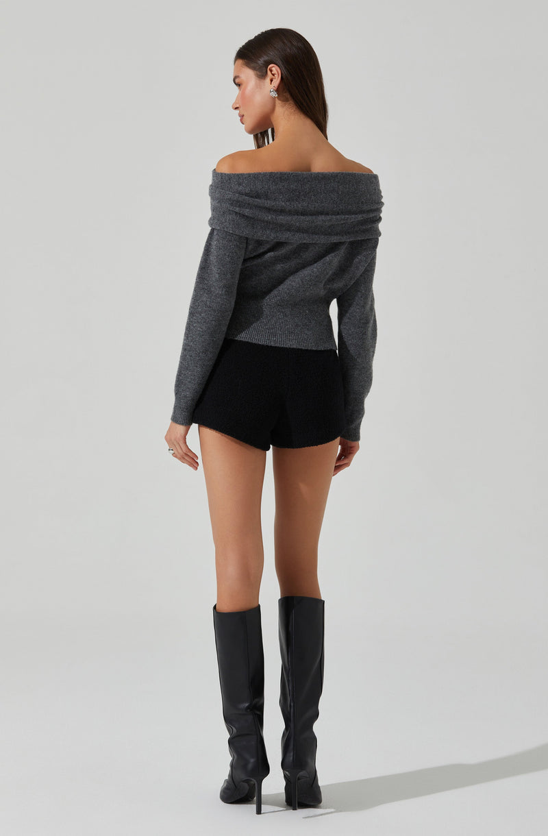 A woman is wearing a wide, off-the-shoulder, folded-over gray sweater with long sleeves. She pairs it with black shorts and tall black high-heeled boots.