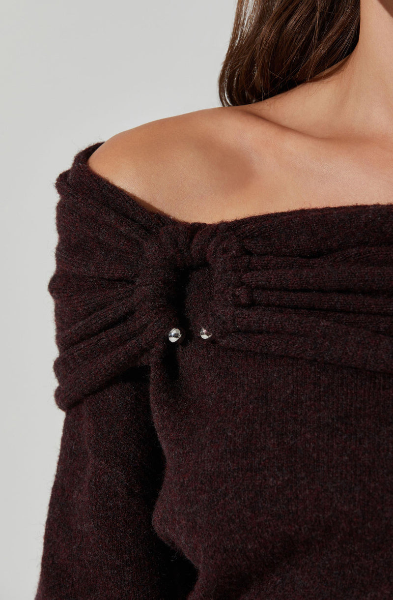 A person is wearing a dark brown, off-the-shoulder sweater with textured, ribbed detailing along the top edge. The sweater has gathered fabric at the shoulder with small decorative metallic beads.