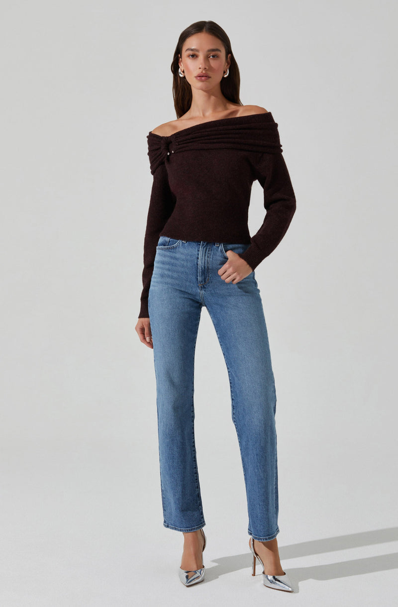 A woman is wearing an off-the-shoulder, long-sleeved, dark brown sweater with a textured, ribbed design across the top. She pairs it with high-waisted, light blue jeans and silver, pointed-toe high heels. Her dark brown hair is styled straight, and she accessorizes with hoop earrings.