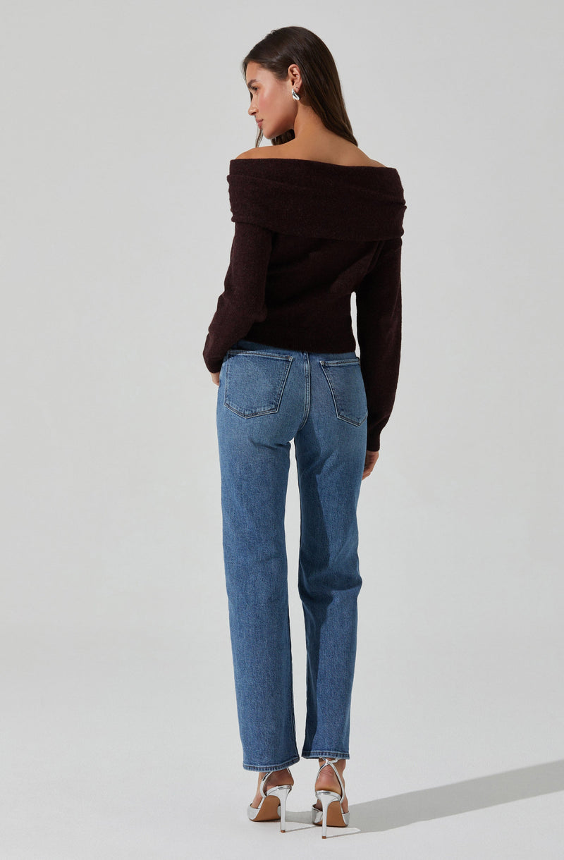 A woman is wearing an off-the-shoulder, long-sleeved sweater in a deep brown color. She is also dressed in high-waisted, straight-leg blue jeans and white high-heeled sandals with ankle straps.