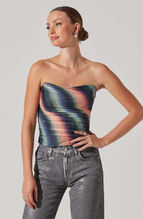A woman is wearing a strapless, multicolored satin top with a pattern of horizontal, blurred stripes in shades of green, pink, blue, and black. She pairs it with high-waisted, metallic gray jeans. She accessorizes with large silver hoop earrings and rings on her fingers.