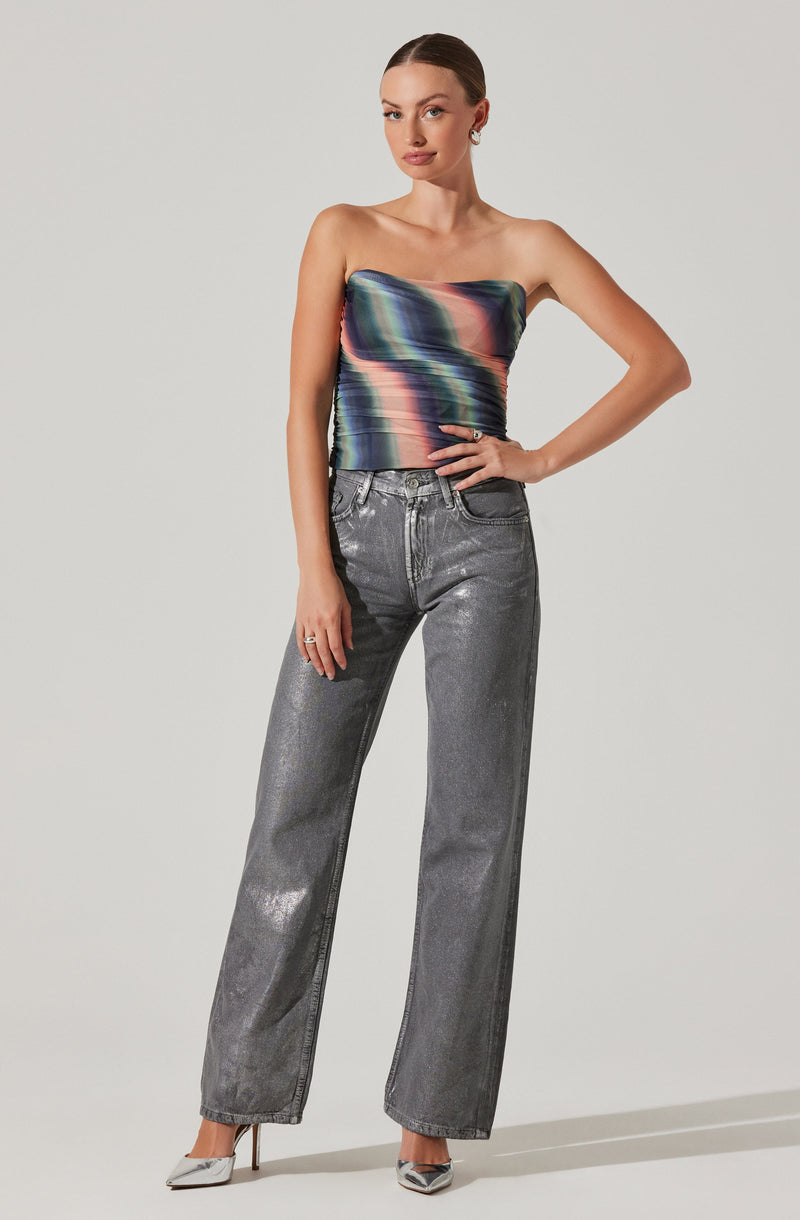A woman is wearing a strapless, multicolored satin top with a shiny, smooth texture and horizontal stripes in shades of green, blue, pink, and black. She pairs it with high-waisted, wide-leg jeans that are metallic silver with a slightly distressed finish. The outfit is complemented with pointed-toe high heels in silver.