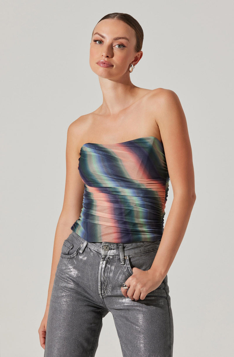 A woman is wearing a strapless, fitted top with a colorful, abstract pattern that features shades of pink, green, blue, and black. The top is ruched along the sides, creating a textured appearance. She pairs it with high-waisted, silver metallic jeans that have a slightly loose fit.