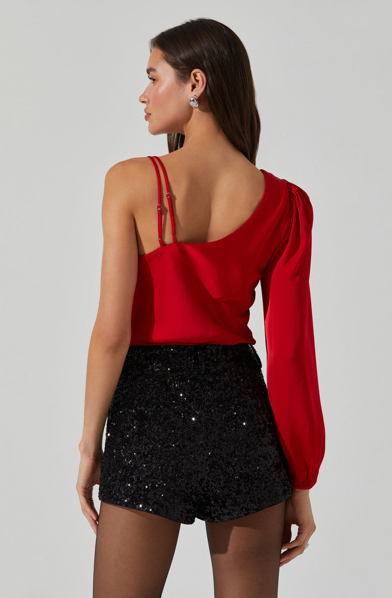 A woman is wearing a red satin top with spaghetti straps on the left shoulder and a long, gathered sleeve on the right arm. She pairs it with high-waisted black shorts that are covered in black sequins, creating a shiny, glittery appearance.