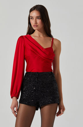 A woman is wearing a red top with a one-shoulder design featuring a draped style on the front, complemented by a single long puffed sleeve. She pairs it with high-waisted black sequined shorts that are fitted and sparkle. The top has thin spaghetti straps on the other shoulder, creating an asymmetrical look.