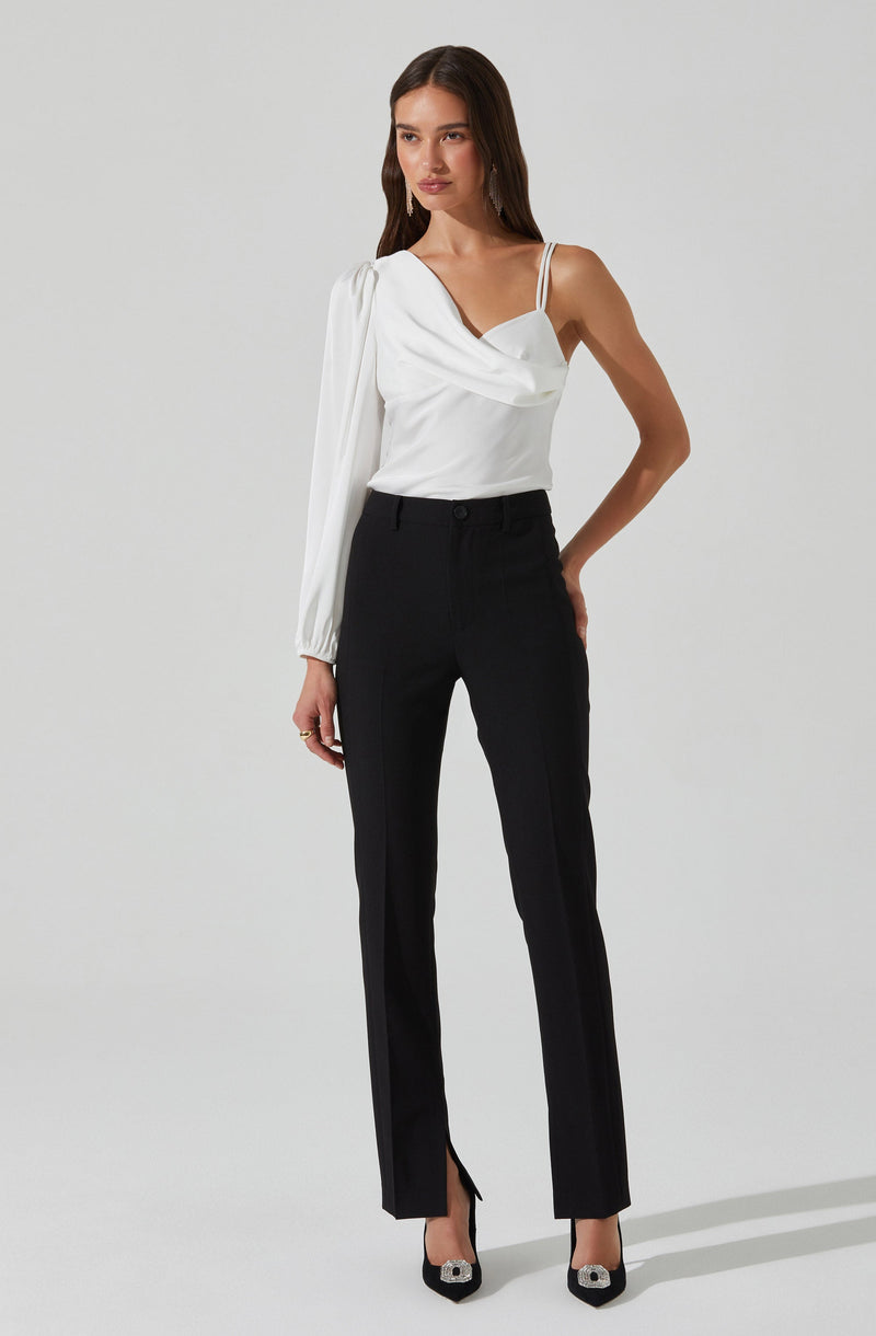 A woman is wearing a white blouse with a draped neckline and spaghetti straps, paired with high-waisted black trousers that have a front slit at the hem. She is also wearing black high-heeled shoes with decorative embellishments.