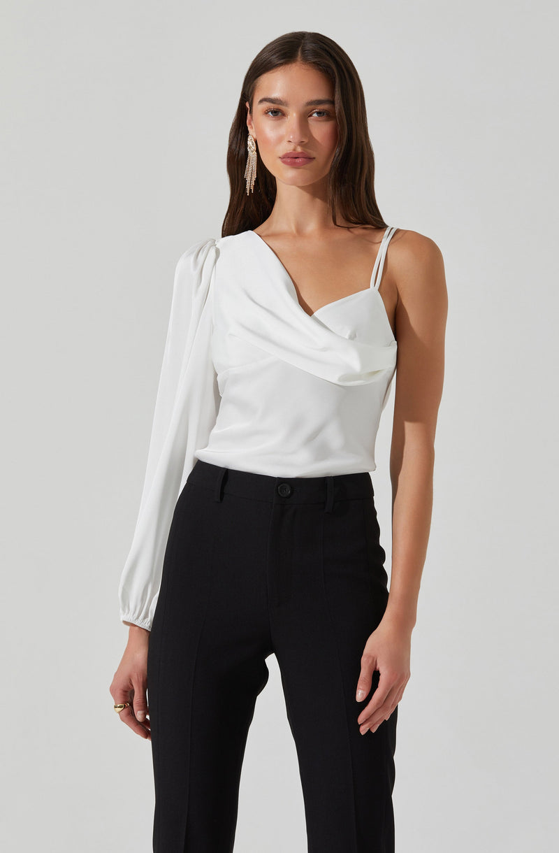 A woman is wearing a white sleeveless top with a draped neckline and one long, puffed sleeve on the left side, while the right side has two thin spaghetti straps. She pairs the top with high-waisted black trousers.