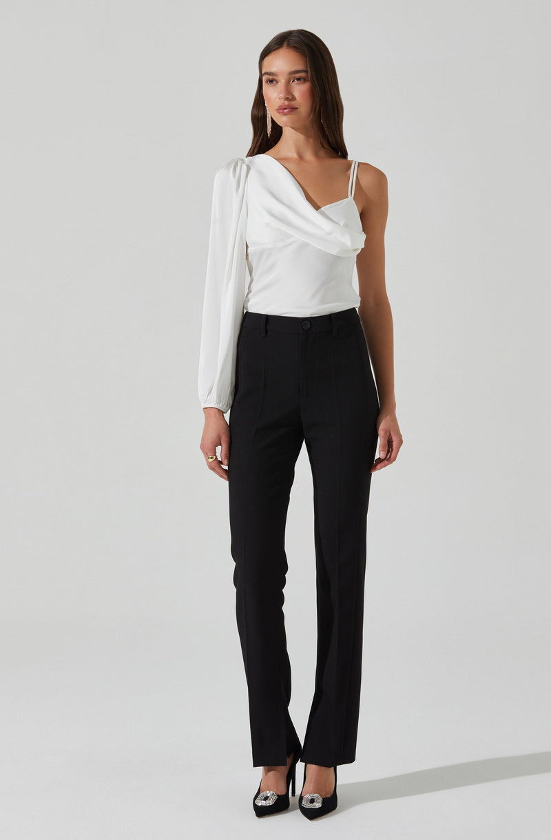 A woman is wearing a white blouse with an asymmetrical, draped design that features a single long sleeve on her left arm and a sleeveless strap on her right shoulder. She pairs it with high-waisted, straight-leg black trousers. She completes her outfit with black pointed-toe heels adorned with decorative embellishments.