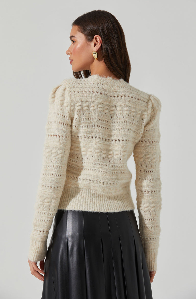 A woman is wearing a cream-colored knitted sweater with a textured open stitch pattern and slightly puffed shoulders, paired with a high-waisted black pleated leather skirt. She has gold hoop earrings and has straight, shoulder-length dark hair.