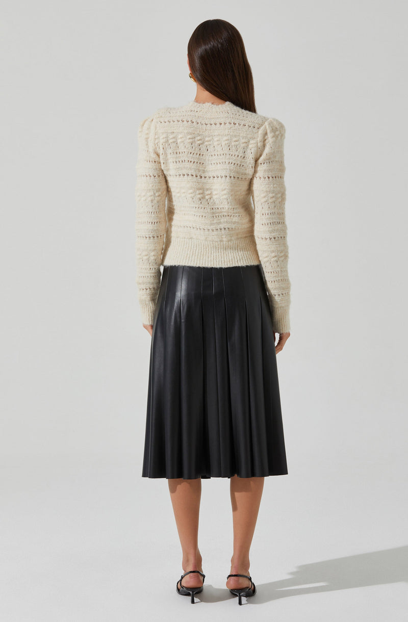 A woman is wearing a cream-colored, long-sleeved, knitted sweater with a textured pattern. She pairs it with a high-waisted, black pleated midi skirt that has a shiny leather appearance. She is also wearing black open-toe, ankle-strap high-heeled shoes.