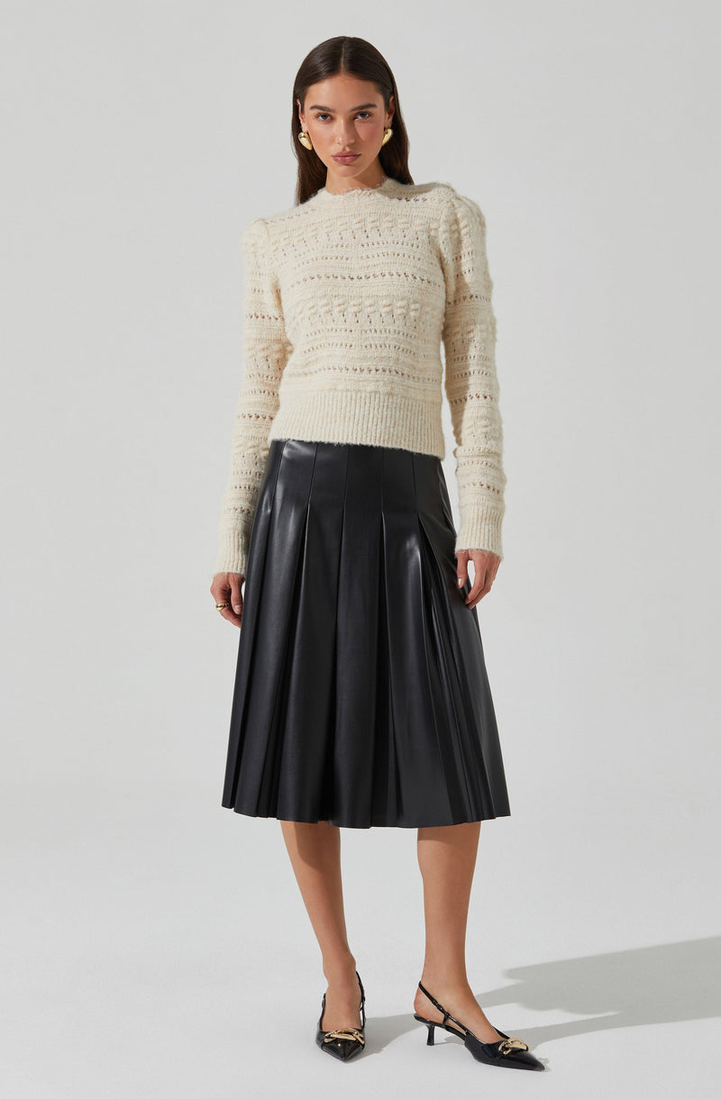A woman is wearing a cream-colored, long-sleeved, knitted sweater with a textured pattern, paired with a black, pleated, midi-length leather skirt. She accessorizes with gold earrings and has on black pointed-toe heels with ankle straps and decorative gold elements.