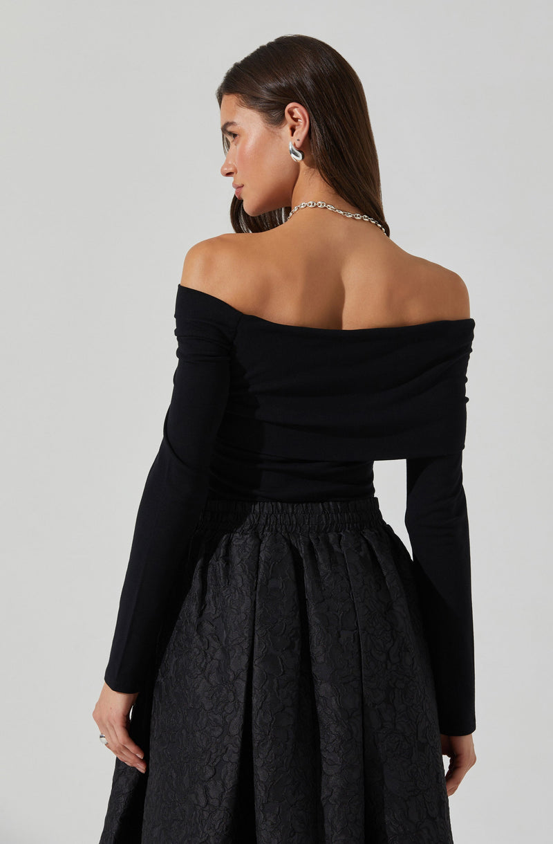 A woman is wearing an off-the-shoulder black top with long sleeves. She pairs it with a high-waisted, textured black skirt that features a subtle floral pattern. She accessorizes with silver hoop earrings and a chain necklace.