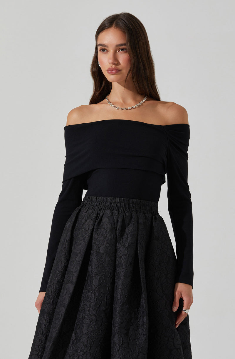A woman is wearing an off-the-shoulder black top with long sleeves and a fitted waistband. She pairs it with a high-waisted, full skirt that has a textured, floral pattern and a gathered, voluminous silhouette. She accessorizes with a silver chain necklace and a ring on her right hand.