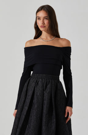 A woman is wearing an off-the-shoulder black top with long sleeves and a fitted waistband. She pairs it with a high-waisted, full skirt that has a textured, floral pattern and a gathered, voluminous silhouette. She accessorizes with a silver chain necklace and a ring on her right hand.