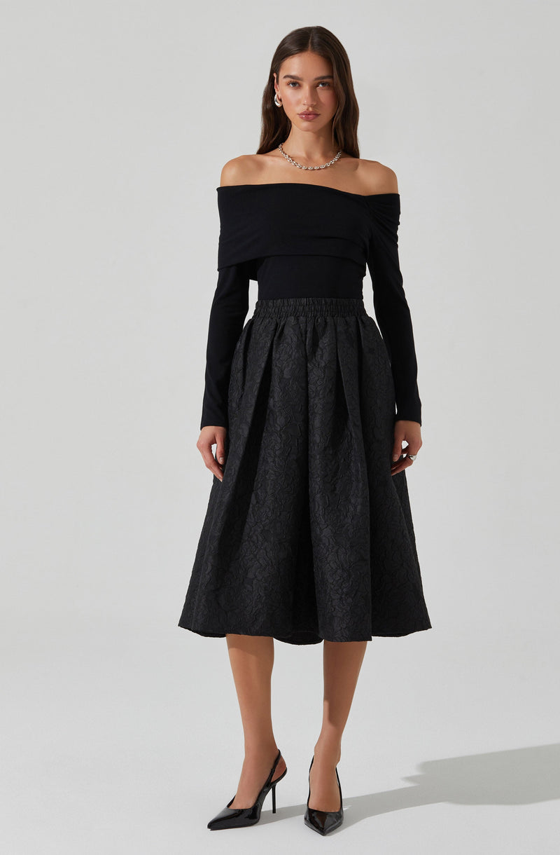 The woman is wearing an off-the-shoulder black top with long sleeves, paired with a high-waisted, full-length black skirt that features a textured, patterned fabric. She accessorizes with a necklace, earrings, and black high-heeled shoes.