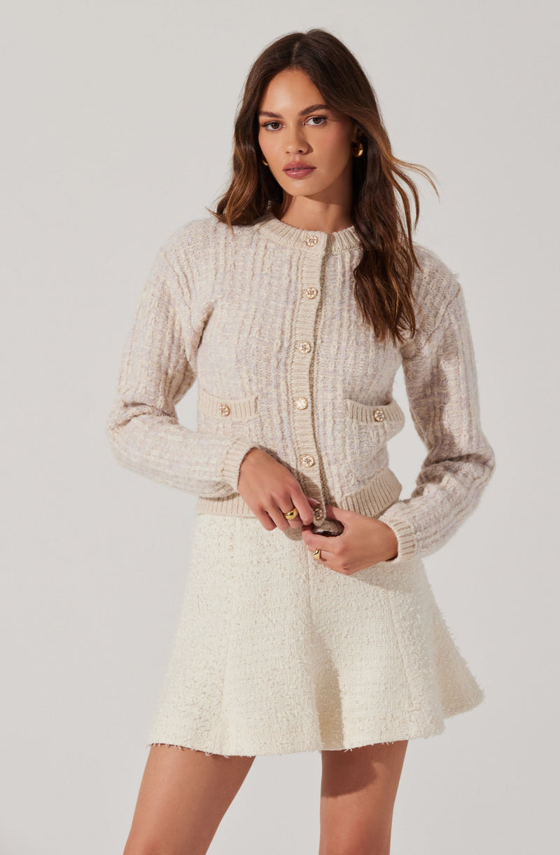 A woman is wearing a light-colored, textured knit cardigan with large buttons down the front, and two small decorative pockets on the front with matching buttons. She has paired it with a cream-colored, textured, A-line mini skirt.