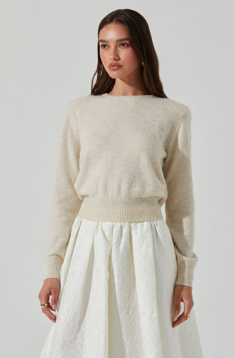 A young woman is wearing a cream-colored, long-sleeved, fuzzy sweater with a crew neckline and ribbed cuffs and hem. She pairs it with an elegant, white, knee-length skirt that has a textured, floral pattern and a gathered waist.