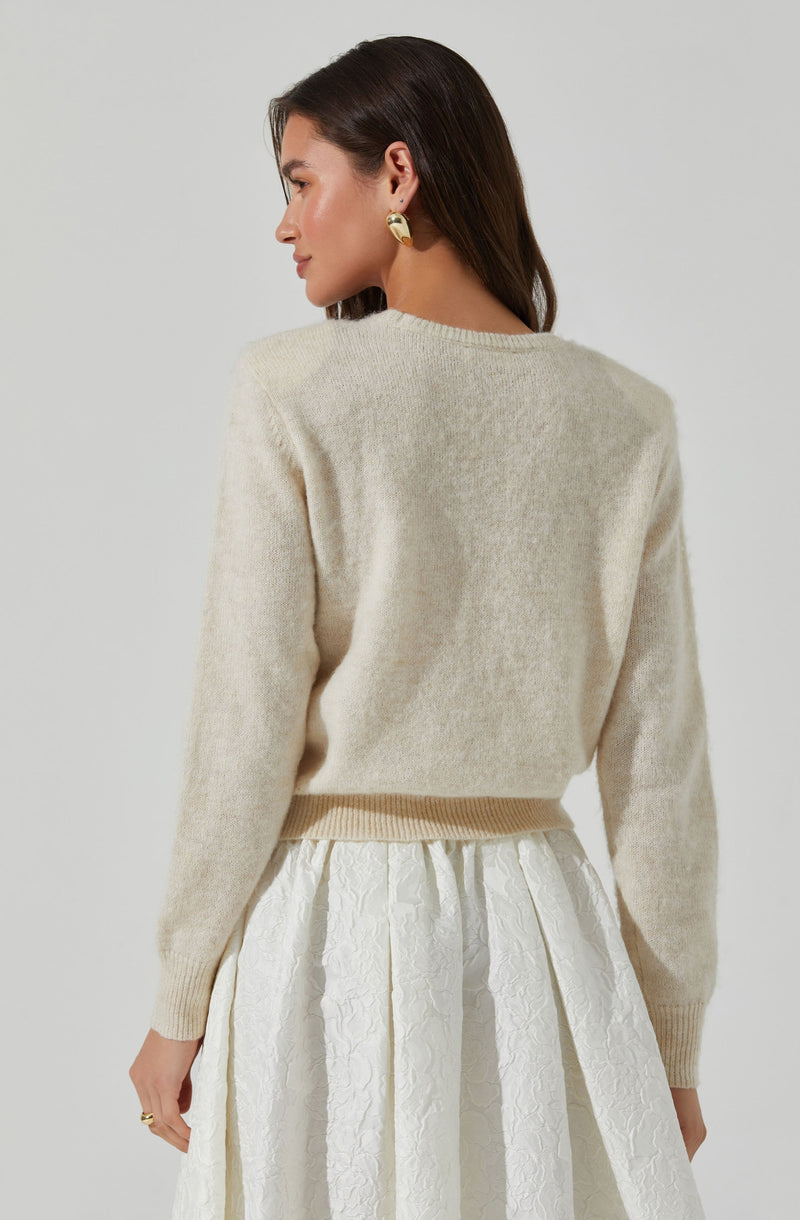 A woman is wearing a cream-colored, long-sleeved, fuzzy knit sweater with a ribbed hem and cuffs. She pairs it with a white, textured, knee-length skirt that features a crinkled fabric and pleats. She has gold hoop earrings and a ring on her right hand.