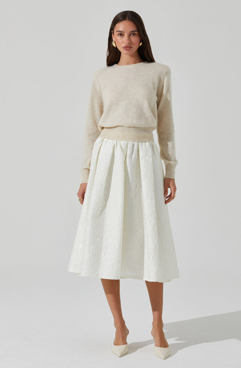 A woman is wearing a light beige, long-sleeve sweater with a crew neckline. The sweater appears soft and fuzzy, with ribbed cuffs and hem. She pairs it with a high-waisted, white, textured midi skirt that has a full, gathered silhouette. She completes the outfit with white pointed-toe heels.