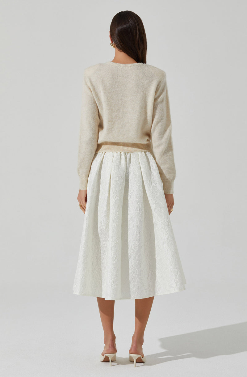 A woman is wearing a light cream, fuzzy knit sweater with long sleeves and a round neckline. She pairs it with a high-waisted, white midi skirt that features a textured, floral pattern and pleats. She completes her outfit with white, open-toe high-heeled shoes with ankle straps.