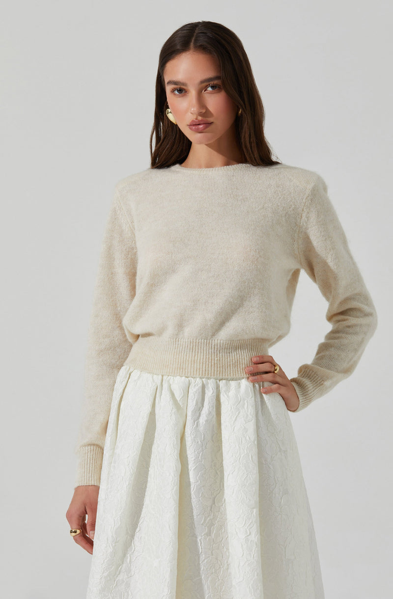 A woman is wearing a soft, cream-colored, knit sweater with a round neckline and long sleeves. She pairs it with a high-waisted, white textured skirt that features pleats and a subtle floral pattern.