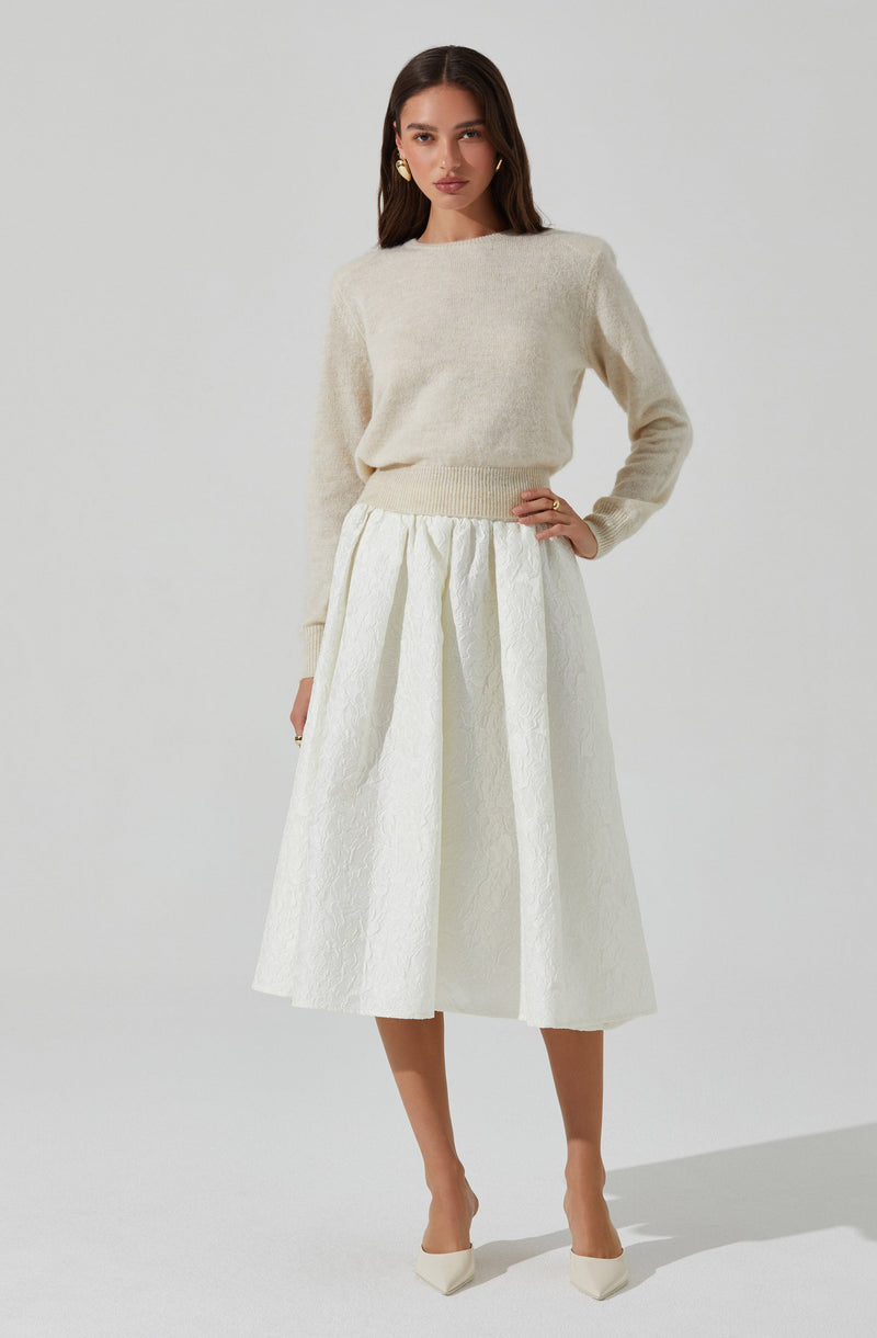 A woman is wearing a soft, cream-colored, long-sleeved sweater with a relaxed fit and a crew neckline. She pairs it with a high-waisted, white, textured midi skirt that features pleats and volume. Her outfit is completed with pointed, cream-colored heels.
