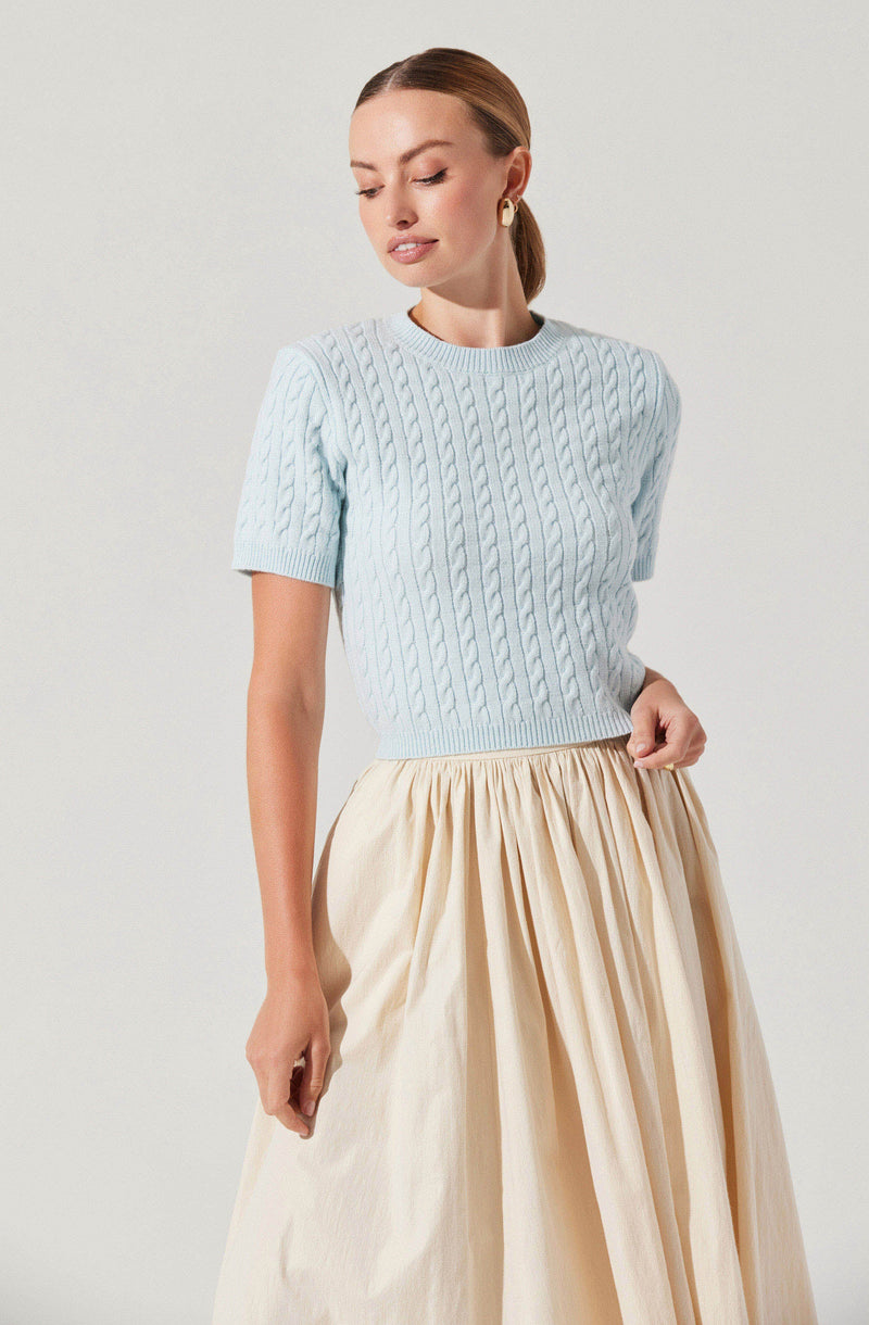 A woman is wearing a light blue, short-sleeved, cable-knit sweater paired with a high-waisted, full beige skirt that has pleats.