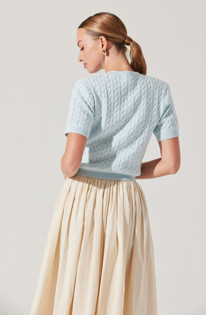 A woman is wearing a light blue, short-sleeved cable-knit sweater and a high-waisted, cream-colored, pleated skirt. The sweater has a textured, woven pattern, and she has her hair tied back in a low ponytail. She is turned slightly to the side with her eyes closed.