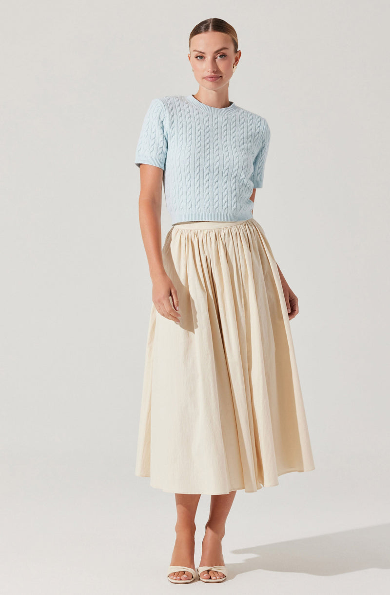 A woman is wearing a light blue, short-sleeved cable-knit sweater paired with a high-waisted, cream-colored, pleated midi skirt. She has beige open-toe heels. Her hair is pulled back in a sleek style, and she is standing against a plain, light background.
