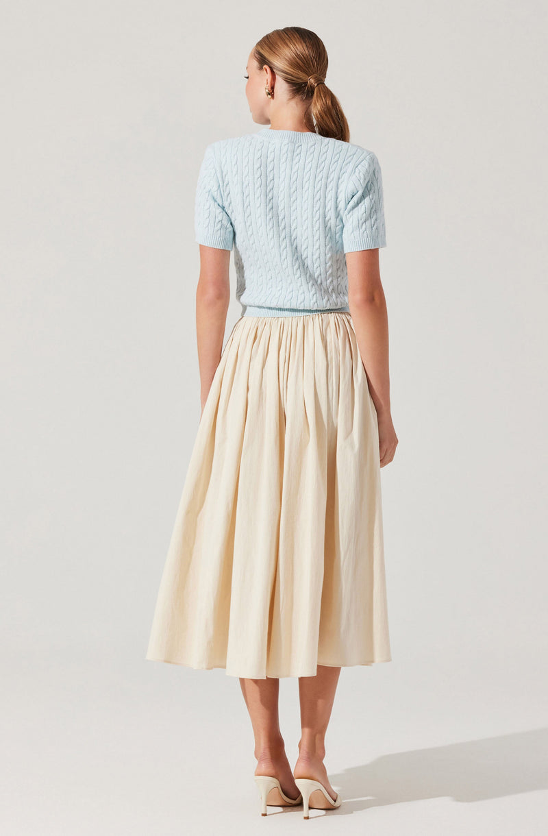 A woman is wearing a light blue, short-sleeved, cable-knit sweater and an ankle-length, cream-colored pleated skirt. She has her hair tied back in a ponytail and is wearing beige high-heeled shoes.