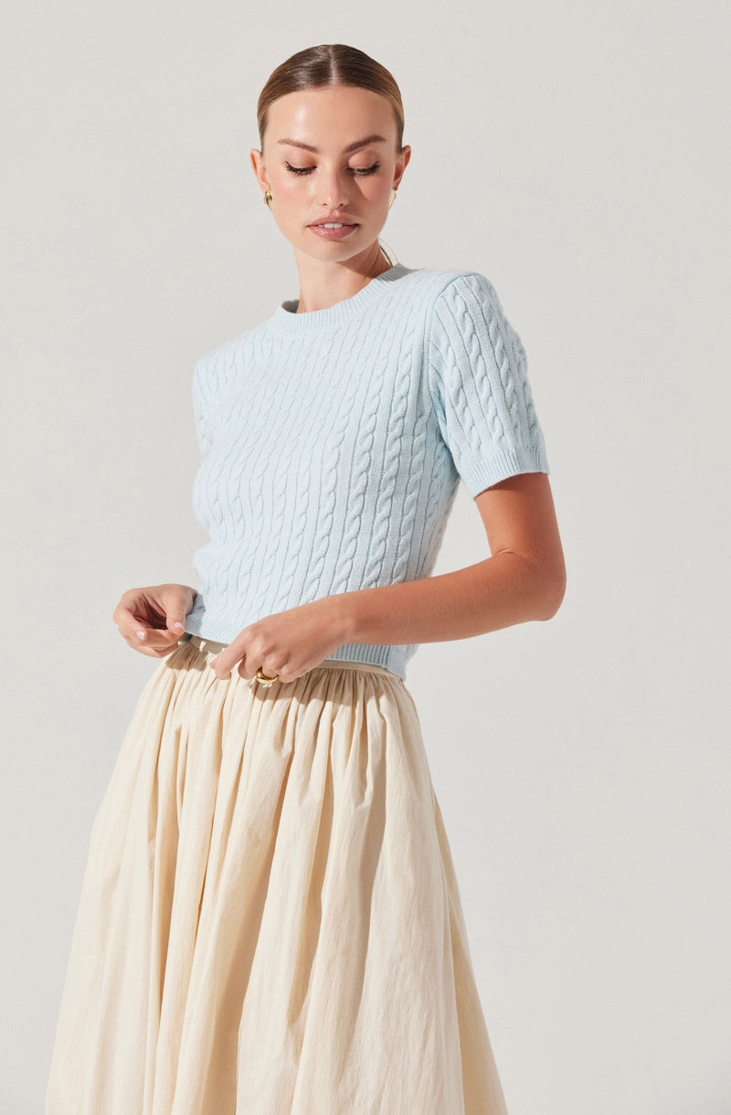 A woman is wearing a light blue short-sleeved cable-knit sweater with a round neckline. She is also wearing a high-waisted, cream-colored, flowy skirt with a gathered waistband.