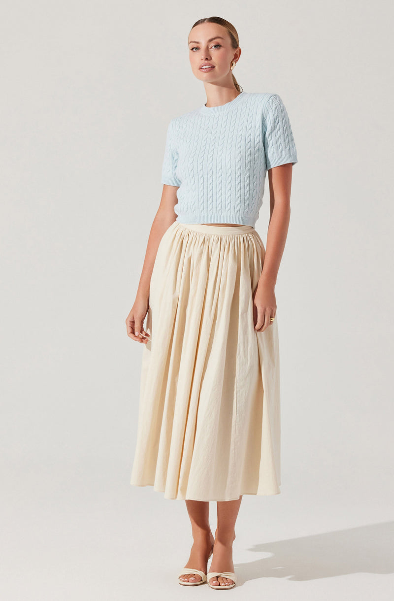 A woman is wearing a light blue, short-sleeved, cable-knit sweater with a round neckline. She pairs it with a high-waisted, full-length, cream-colored skirt that has a gathered, flowing design. She completes the outfit with open-toe, cream-colored heels.