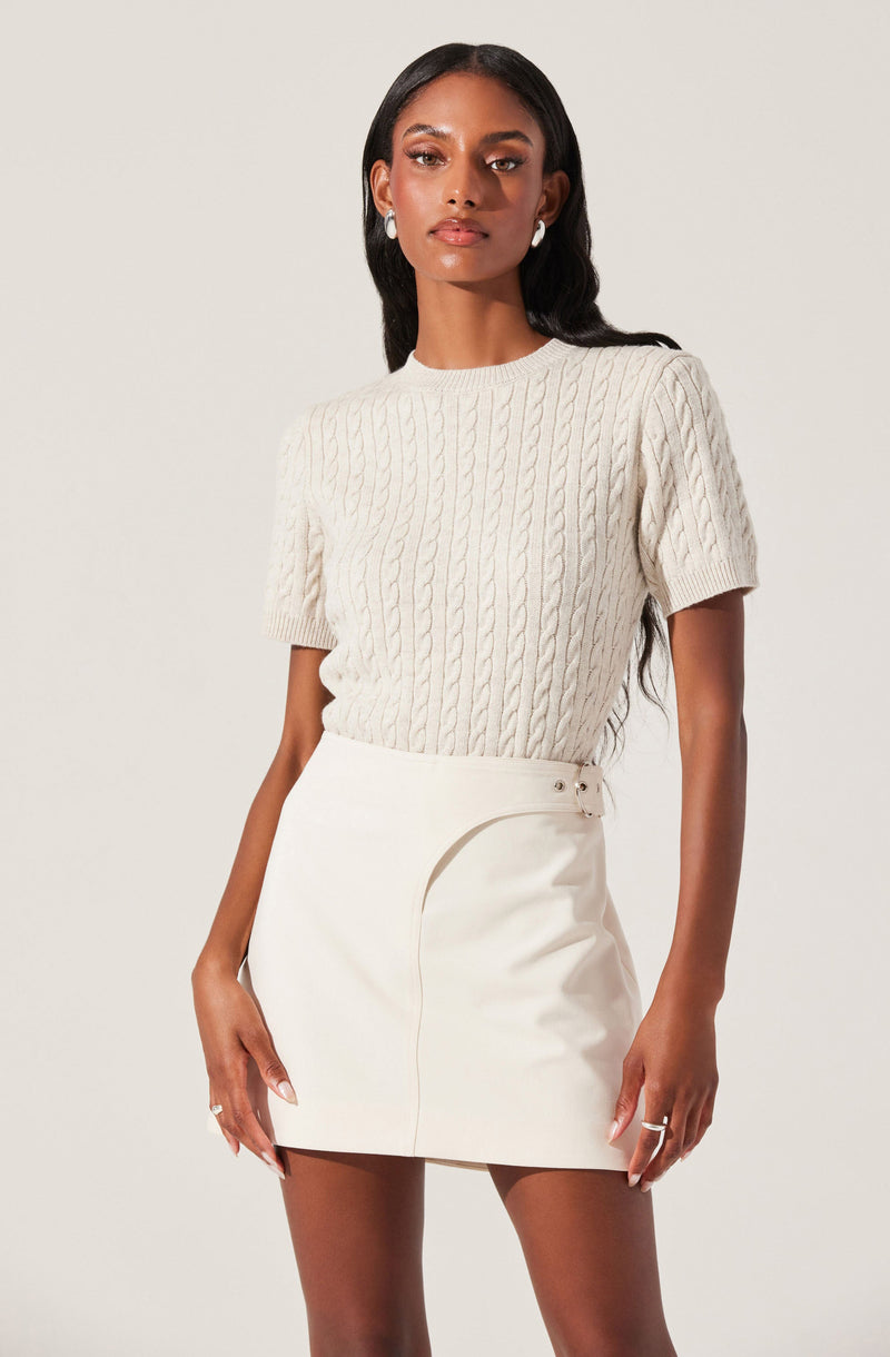 A woman is wearing a short-sleeved, cable-knit cream sweater and a high-waisted, cream skirt with a side slit and buckle detail.