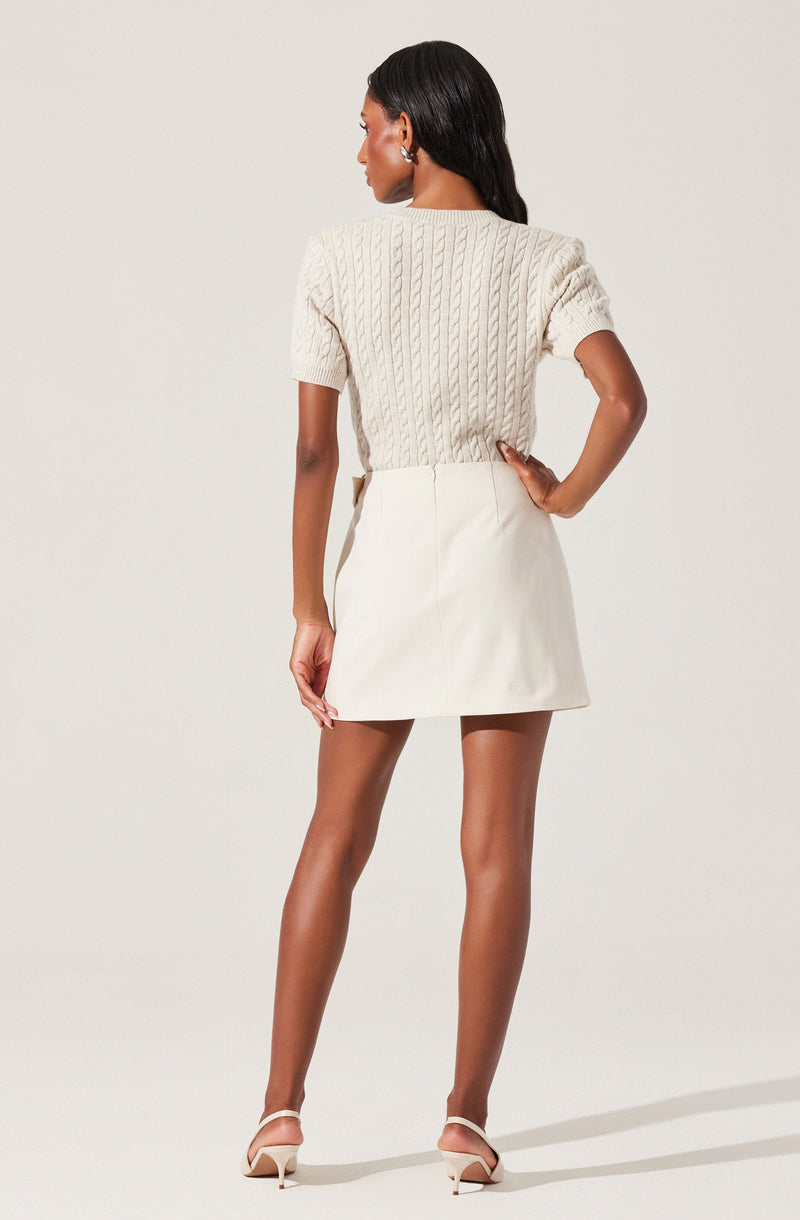 A woman is wearing a cream-colored, short-sleeved, cable-knit sweater paired with a matching cream-colored A-line mini skirt. She is also wearing nude high-heeled sandals.