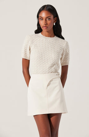 A woman wearing a cream-colored, short-sleeved cable knit sweater with a round neckline. She has on a high-waisted, cream-colored skirt with an asymmetrical zipper detail on the side. She accessorizes with large pearl earrings.