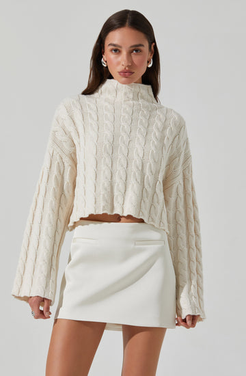 A woman is wearing an oversized, cream-colored cable-knit sweater with a high neckline and long, wide sleeves. She pairs the sweater with a high-waisted, white mini skirt that has a smooth, structured design. She accessorizes with large, white hoop earrings and has long, straight brown hair.