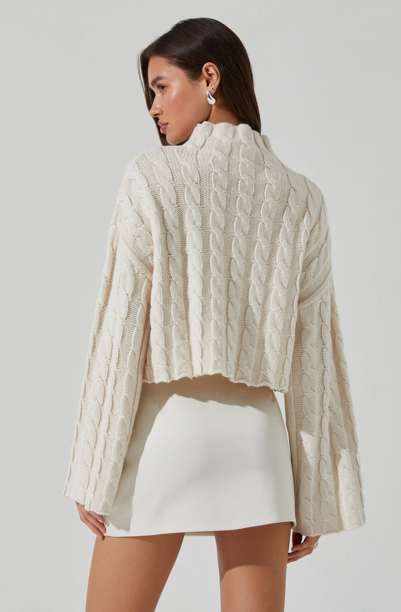 A woman is wearing a cream-colored, cable-knit cropped sweater with a high neckline and long, wide sleeves. She pairs it with a white skirt that has a smooth, fitted appearance.