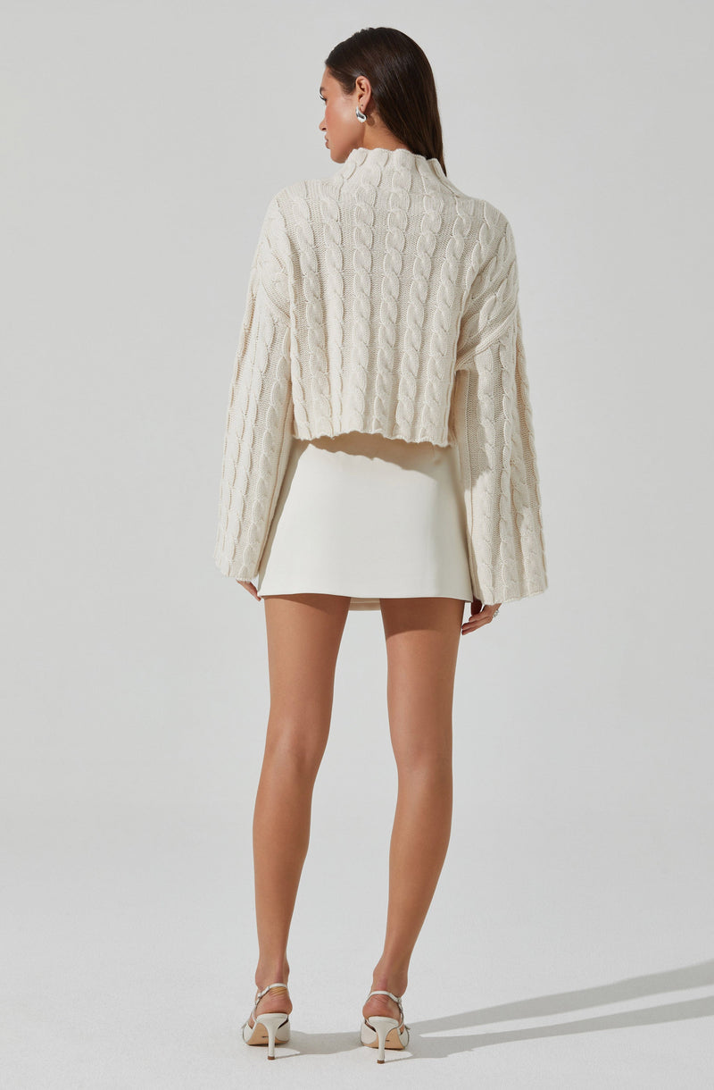 A woman is wearing a cream-colored, loose-fitting, cable-knit sweater with long, wide sleeves. She pairs it with a short, fitted white skirt and white high-heeled shoes.