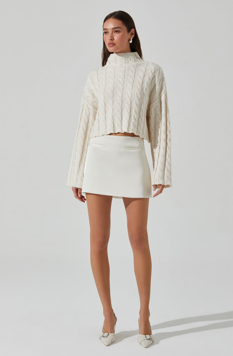 A woman is wearing an oversized, cream-colored, cable-knit sweater with a high neckline and long sleeves that extend past her wrists. She pairs it with a high-waisted, white, mini skirt that has a subtle pocket detail on the front. She completes the outfit with pointed-toe heels that have bow accents.