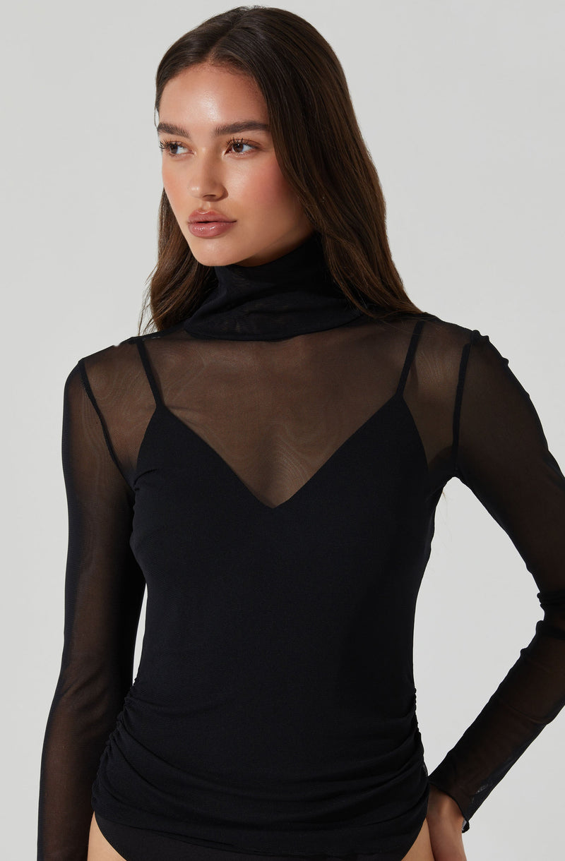 A woman is wearing a black top with a sheer, long-sleeved overlay. The overlay has a high turtleneck collar and is made of a see-through, lightweight fabric. The top underneath has a V-shaped neckline. The shirt fits snugly and is gathered slightly at the waist.