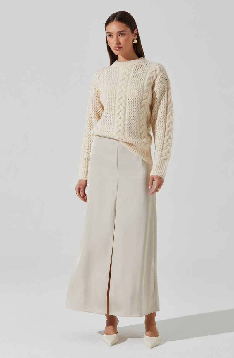 A woman is wearing a cream-colored, chunky cable-knit sweater featuring a variety of textured knit patterns, including large cable designs and smaller stitches. The sweater has a crew neckline and long sleeves. She pairs it with a high-waisted, floor-length cream skirt with a front slit that reaches up to her calves. She completes her look with cream-colored pointed-toe shoes and gold earrings.
