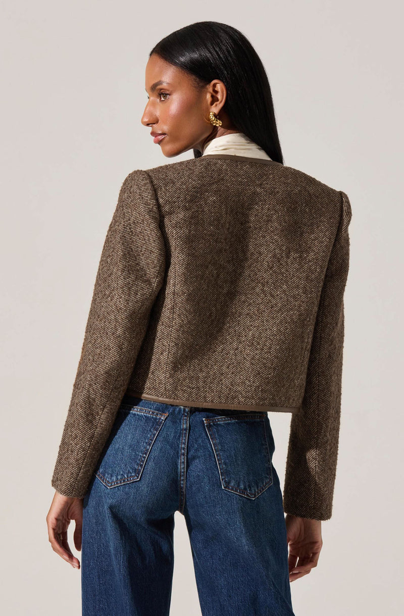 A woman is wearing a brown, textured, cropped blazer with slightly padded shoulders, paired with dark blue high-waisted jeans. The blazer has a subtle herringbone pattern and a structured fit. She also has on a white shirt underneath the blazer and gold earrings.