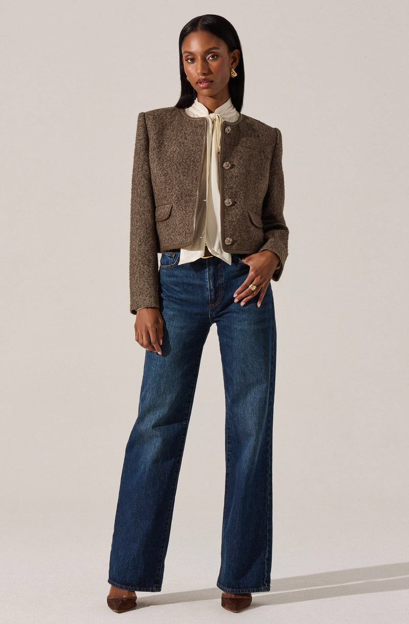 A woman is wearing a cropped, structured blazer in a textured brown fabric with a collarless neckline and three buttons down the front. Underneath, she has a light-colored blouse with a tie detail at the collar. She pairs the top with high-waisted, wide-leg blue jeans and brown pointed-toe shoes. She accessorizes with gold earrings and rings.